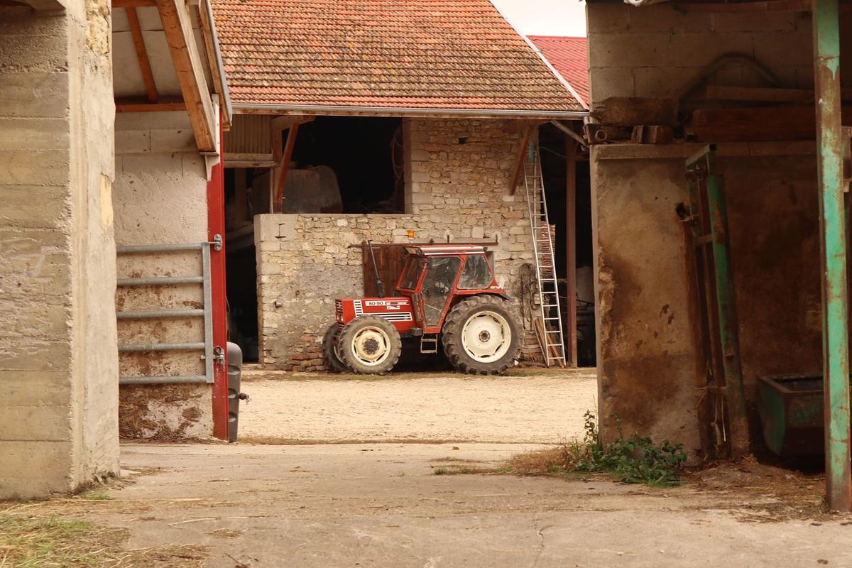 Tracteur rouge garé dans une cour de ferme, entre des bâtiments en pierre et des hangars